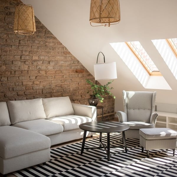 Cozy attic living room with brick accent wall, beige sectional sofa, patterned armchair, and skylight windows.