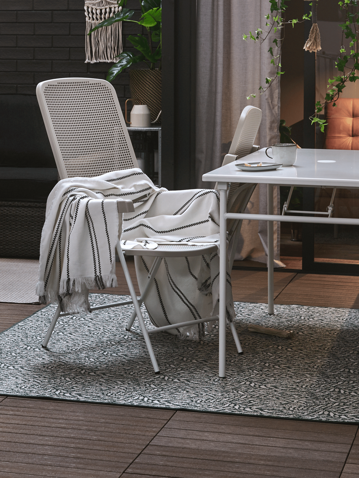 Cozy balcony setup with white metal chairs and a table, soft textiles, and potted plants around.