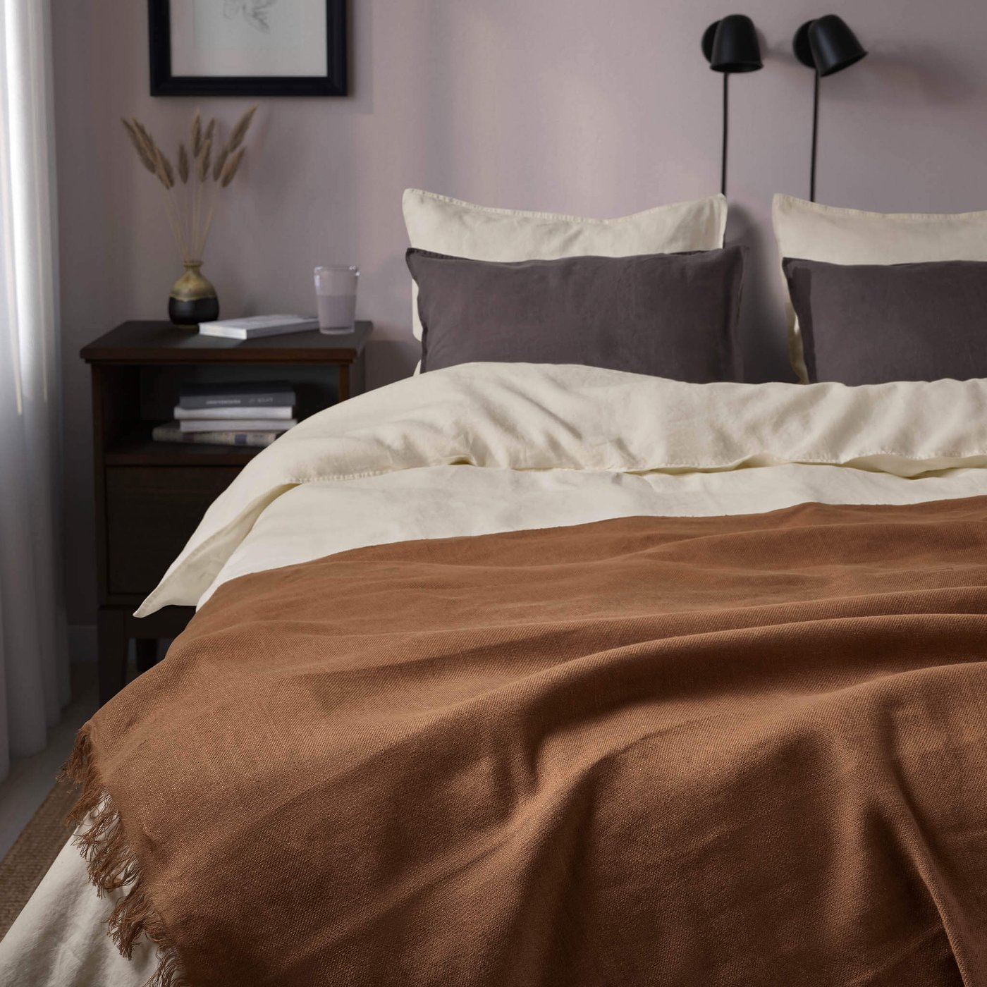 Cozy bed with cream sheets, dark gray pillows, and a brown throw blanket in a softly lit bedroom.