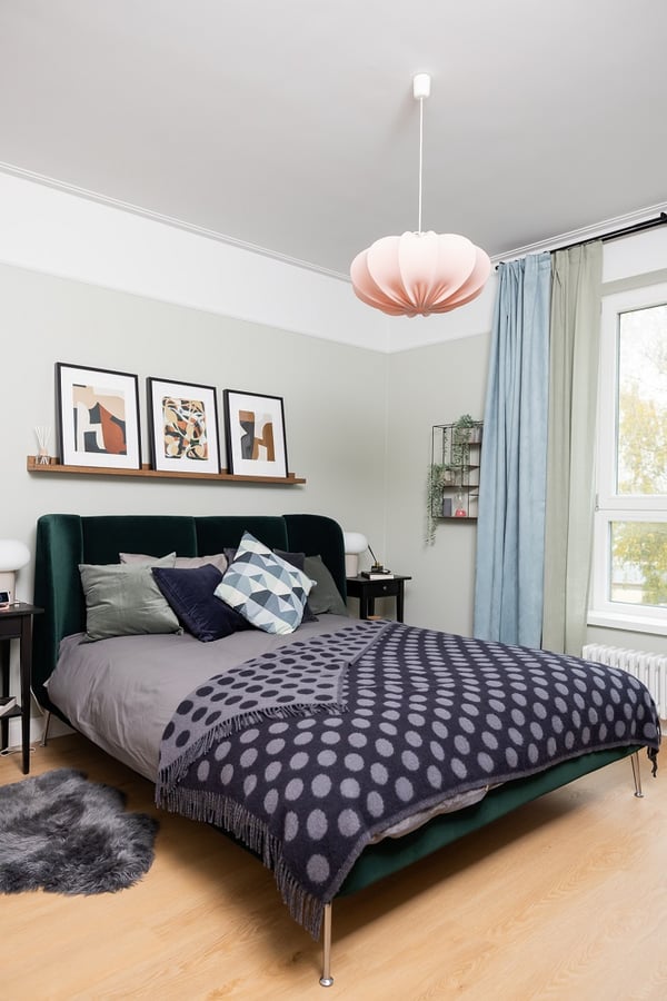 Cozy bedroom with green velvet bed frame, patterned bedding, abstract wall art, and pastel curtains.