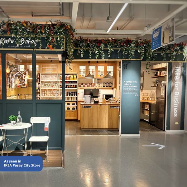 Cozy cafe setup inside an IKEA store, featuring wood accents, lush greenery above, and a small table with chairs in front. Inviting and modern ambiance.