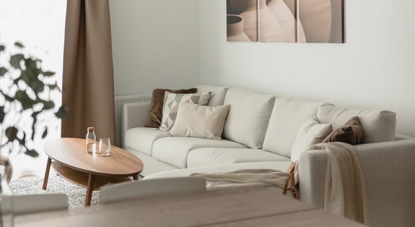 Cozy living room with a large light beige sofa decorated with soft neutral pillows and a brown throw. A wooden oval coffee table with water bottles and glasses sits on a light rug. The room is softly lit by natural light coming through sheer curtains and a warm-toned curtain on the side. A minimalistic abstract artwork hangs above the sofa.