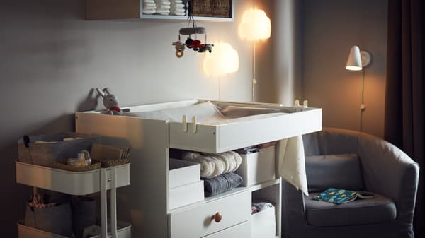 Cozy room with a changing table, a storage trolley with baby accessories, an armchair, night wall lamps and wall storage.