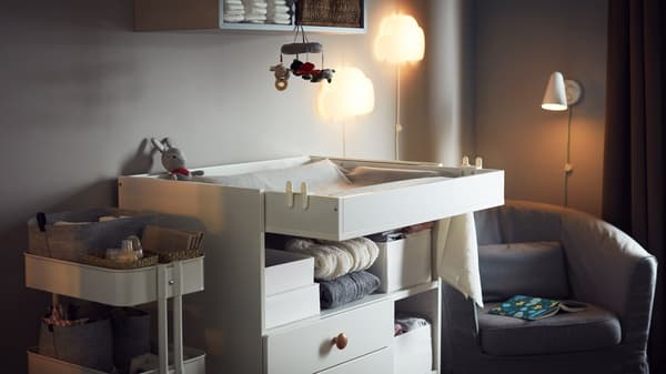 Cozy room with a changing table, a storage trolley with baby accessories, an armchair, night wall lamps and wall storage.