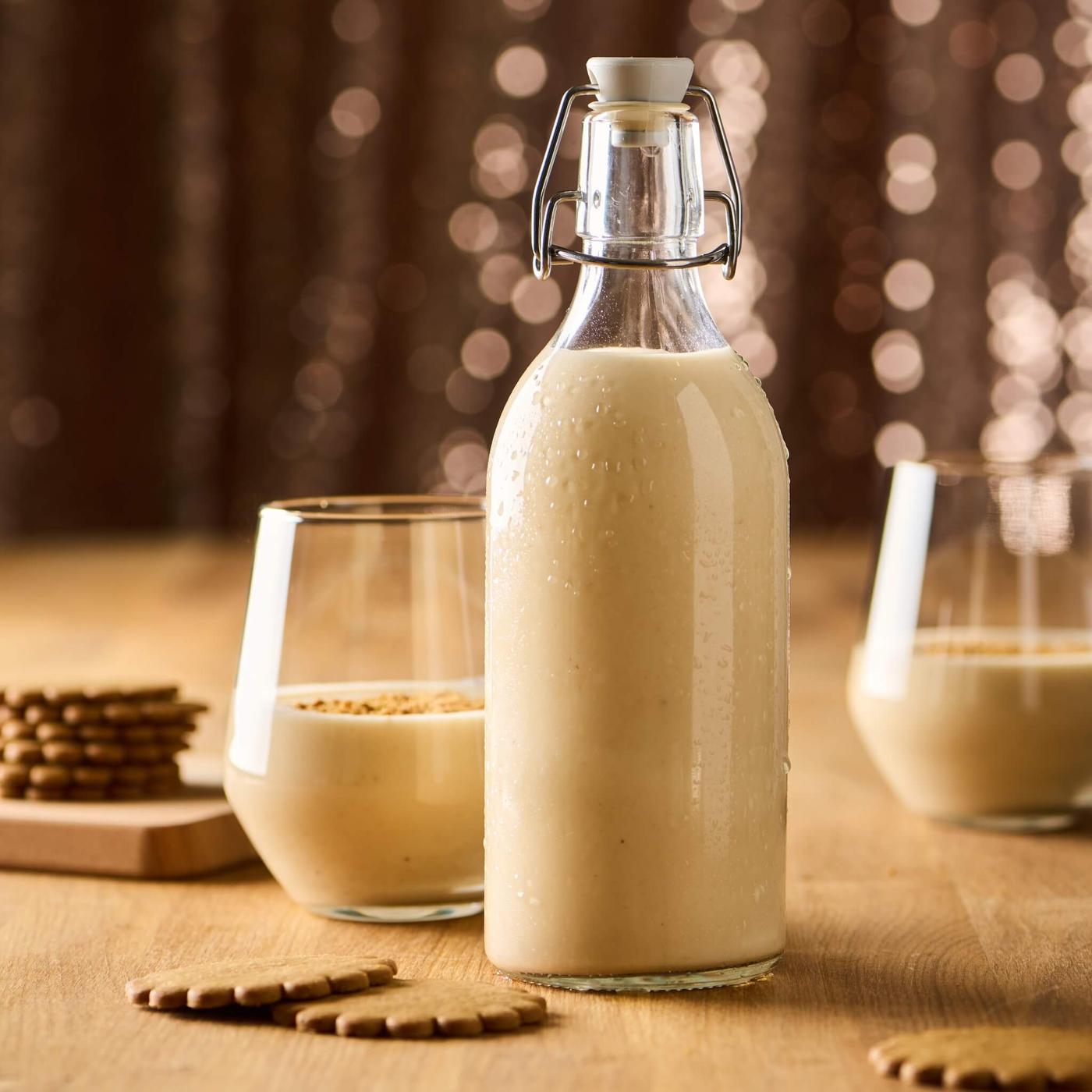 Creamy coquito in a glass bottle on a wooden table, with two filled glasses and cookies arranged around it.
