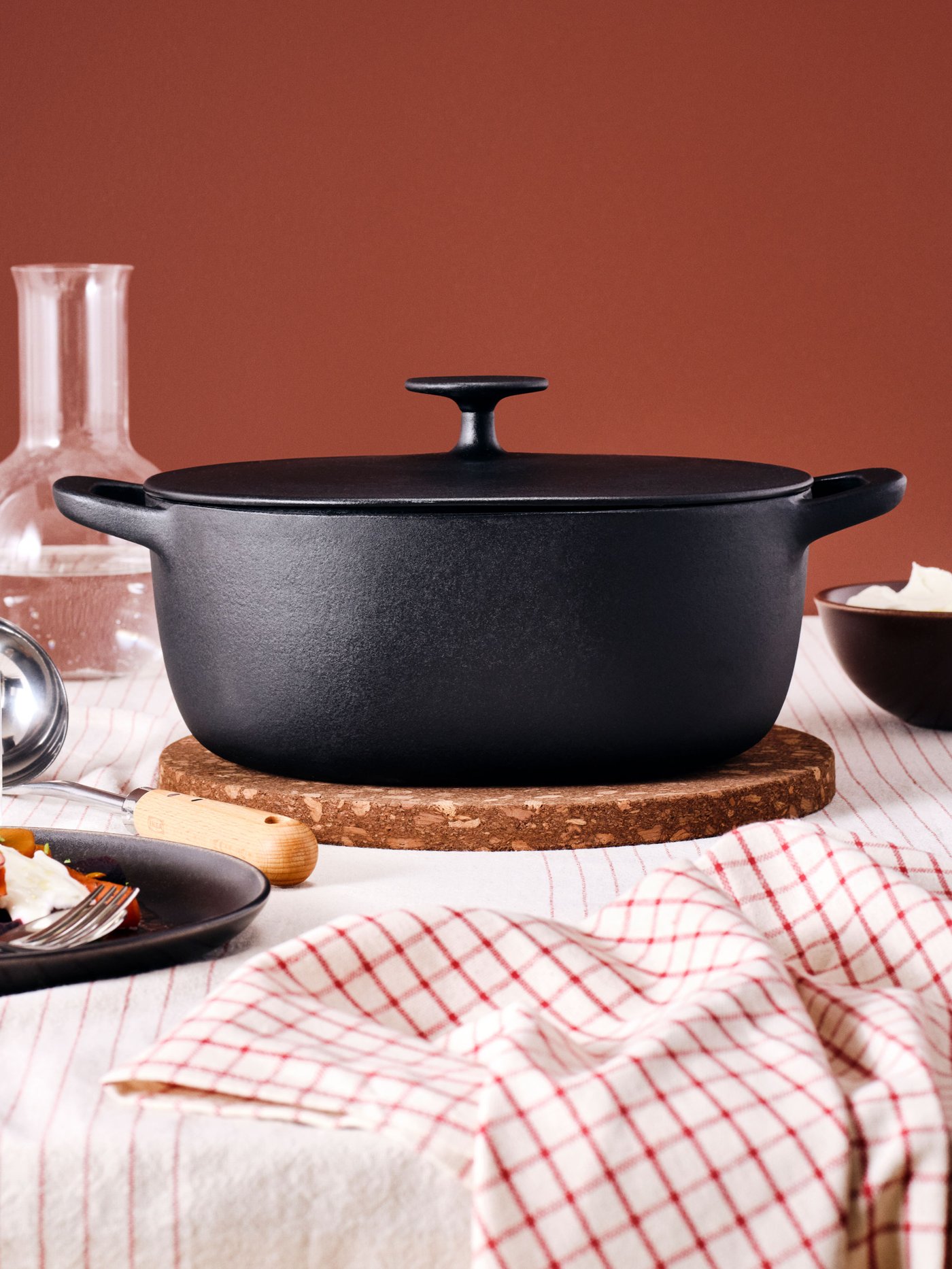 Crni lijevani lonac s poklopcem na okruglom podlošku od pluta, okružen kuhinjskim priborom: staklenom karafom, zdjelicom, metalnom žlicom i kuhinjskom krpom s crvenim kvadratićima.