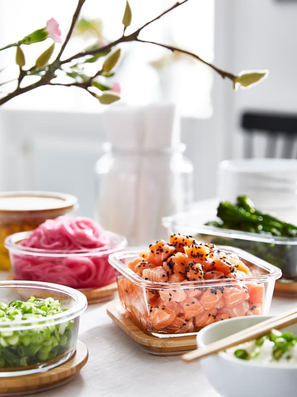 Cuatro recipientes de vidrio para alimentos IKEA 365+, llenos de varios alimentos preparados, se encuentran sobre sus tapas de bambú detrás de un tazón de arroz.