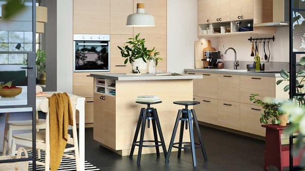 Cucina in rovere chiaro con alcuni sgabelli da bar e piante intorno. Sopra c'è una lampada bianca 