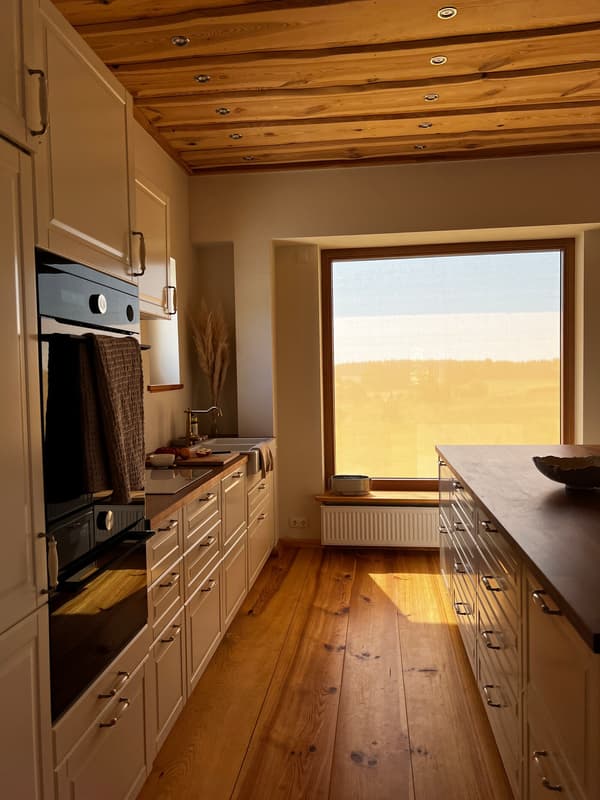 Cucina moderna con soffitto in legno, armadi semplici e luce naturale che entra da una grande finestra, con pavimento in legno caldo.