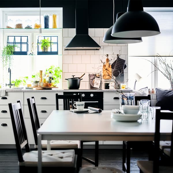 Cuisine VEDDINGE blanche avec cinq chaises STEFAN brun-noir autour de tables MELLTORP blanches, sous des suspensions SKURUP noires.