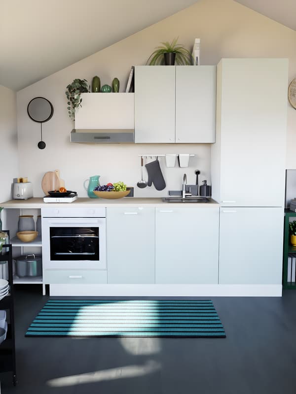 Cuisine verte claire sous pente, avec plan de travail en bois, sol noir, tapis vert. Un réfrigérateur encastré sur la droite, un four et une hotte sur la gauche du meuble de cuisine. 