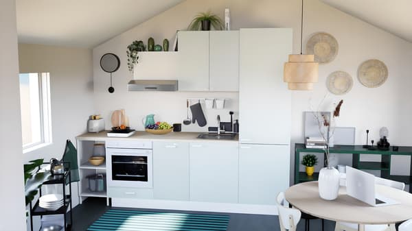 Cuisine verte claire sous pente, avec plan de travail en bois, sol noir et linge de cuisine vert. Abat-jour en osier, et table à manger ronde au premier plan, avec ordinateur et décorations.