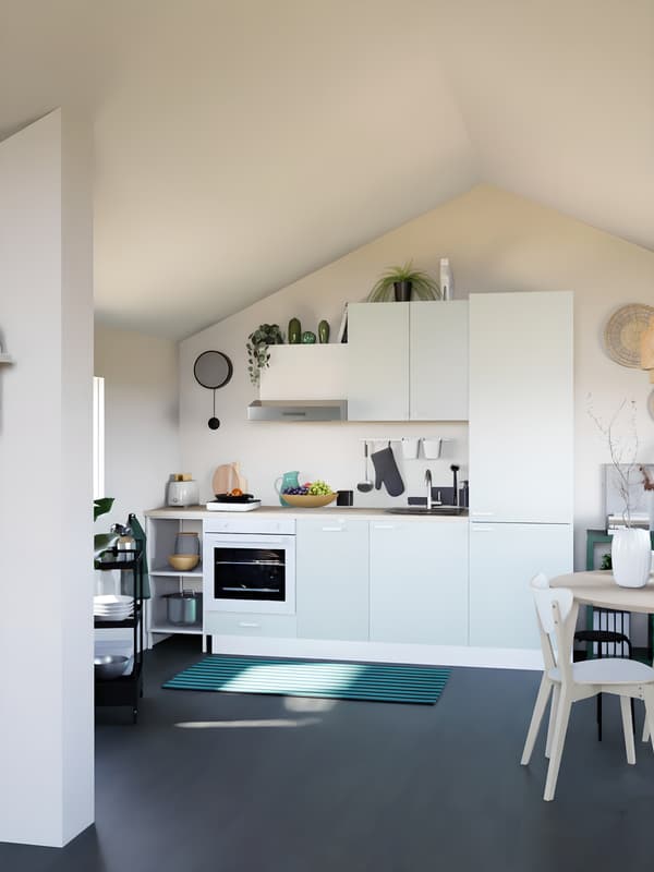 Cuisine verte claire sous pente, avec plan de travail en bois, sol noir et linge de cuisine vert. Abat-jour en osier, et table à manger ronde au premier plan, avec ordinateur et décorations.