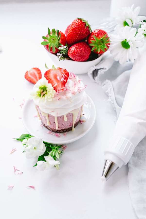 Cupcake mit Sahne und Erdbeere auf weissem Teller, Schale mit Erdbeeren, Blumen und Spritzbeutel im Hintergrund.