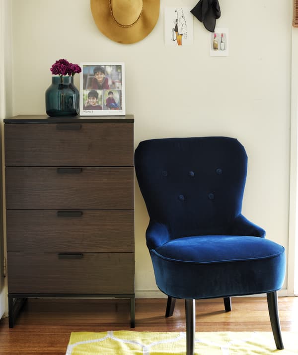 Curruncho dun dormitorio branco cunha cómoda de madeira escura e unha cadeira de brazos de veludo azul escuro.