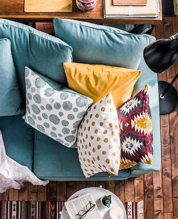Cuscini colorati in diverse fantasie e geometrie su un divano visto dall'alto. Accanto, un tavolino GLADOM con un libro, una candela accesa e degli occhiali.