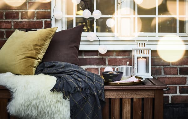 Cushions and throws, a tray with a bowl and a cup, and a lantern on a bench outside a window hung with a lighting chain.