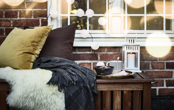 Cushions and throws and a tray with a bowl, a cup and a lantern on a bench outside a window decorated with fairy lights.