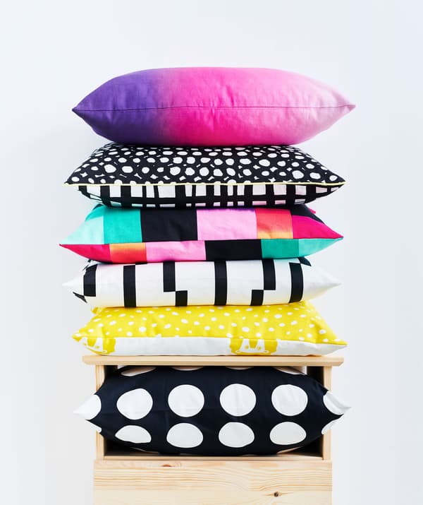 Cushions – black and white, graphic patterns, sharp coloring in a mix – piled in a stack on top of  a chest of drawers.