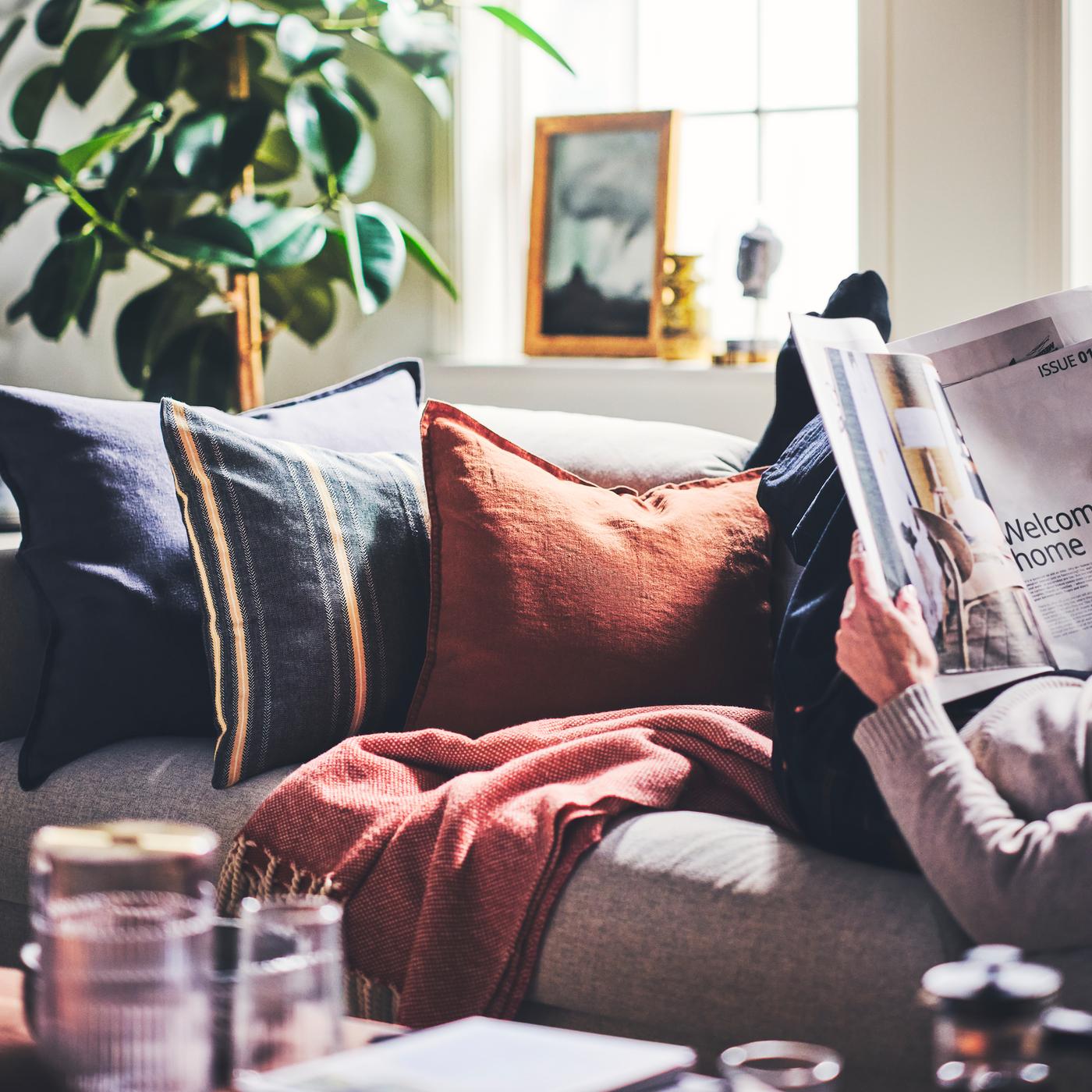 Cushions dressed in a STORTIMJAN cushion cover and DYTÅG cushion covers placed on a sofa next to a person reading a magazine.