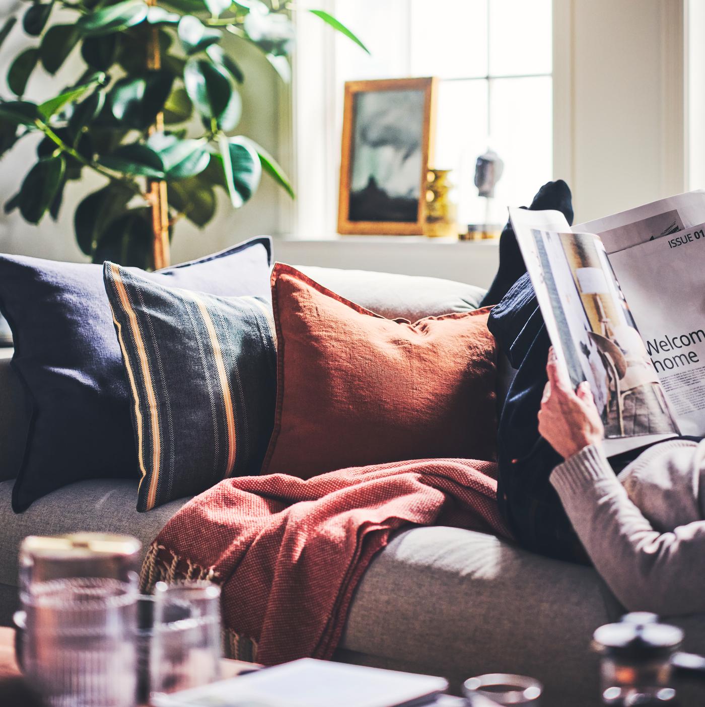 Cushions dressed in a STORTIMJAN cushion cover and DYTÅG cushion covers placed on a sofa next to a person reading a magazine.