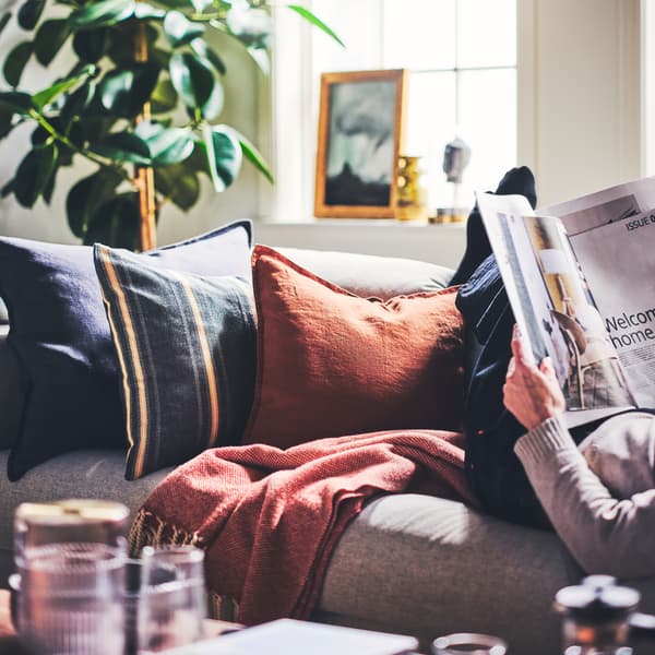 Cushions dressed in a STORTIMJAN cushion cover and DYTÅG cushion covers placed on a sofa next to a person reading a magazine.