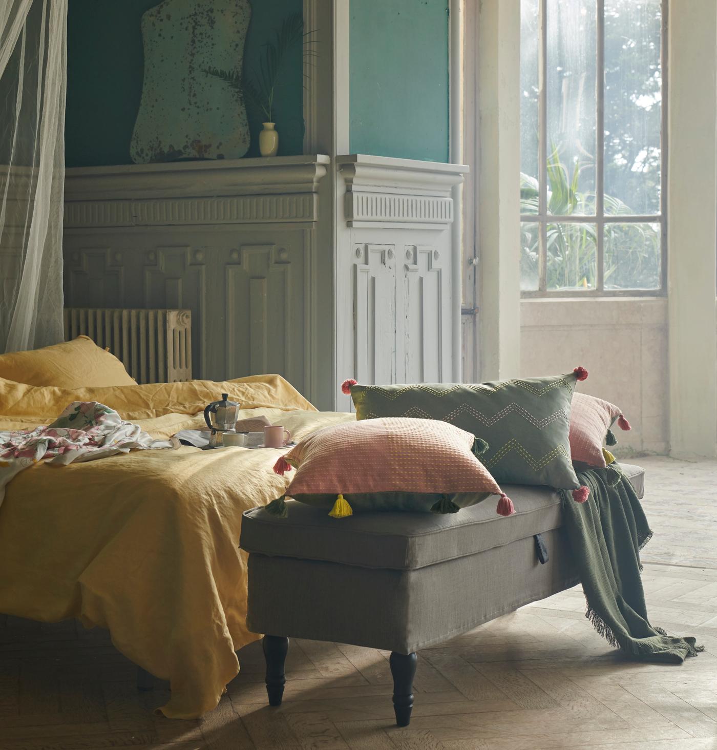 Cushions in KLARAFINA and MOAKAJSA covers lie on a STOCKSUND bench at the foot of a bed with yellow PUDERVIVA bed linen.