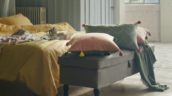 Cushions in KLARAFINA and MOAKAJSA covers lie on a STOCKSUND bench at the foot of a bed with yellow PUDERVIVA bed linen.