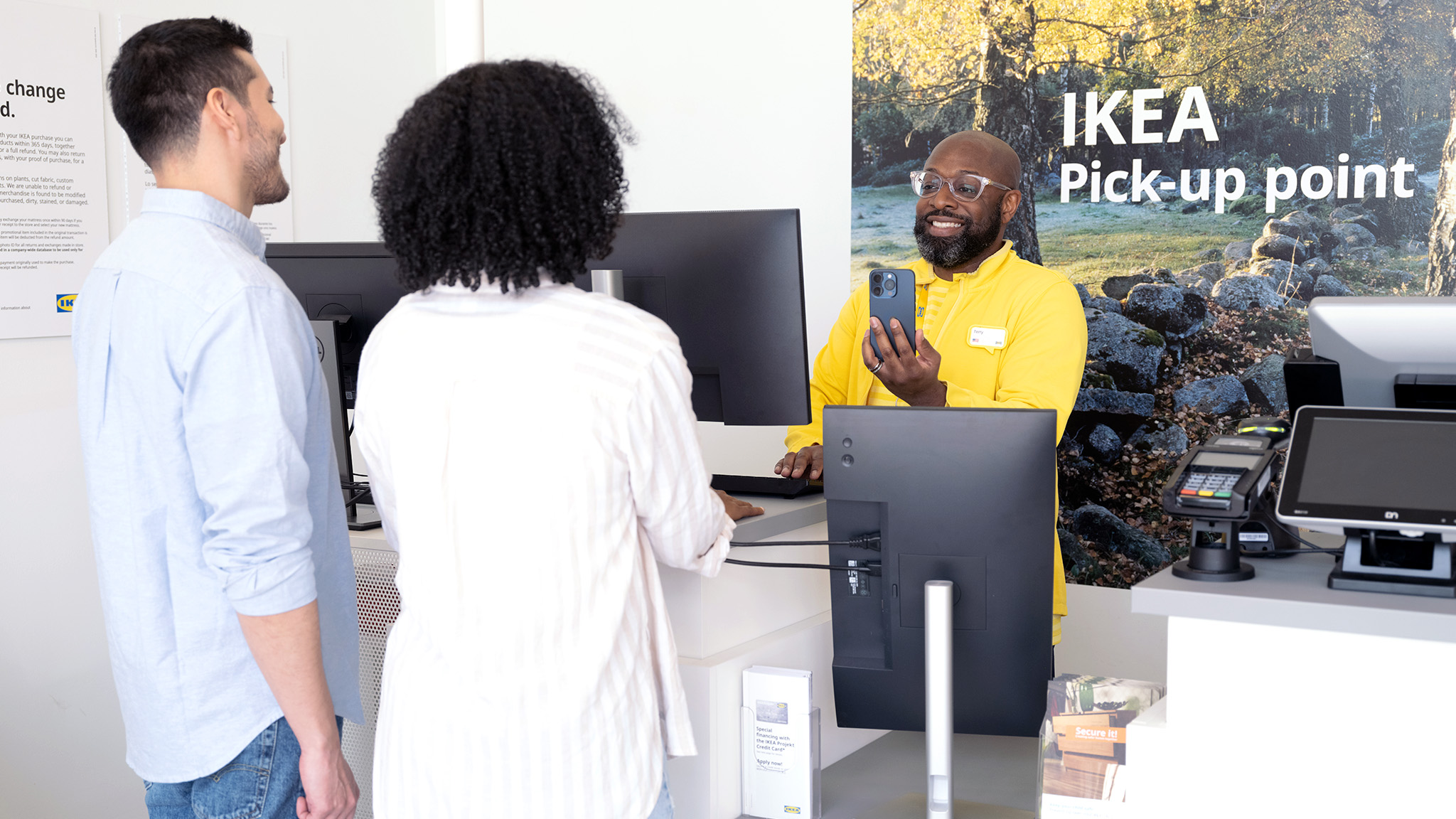 Customers at an IKEA pick-up point counter speaking with an employee holding a phone near computer monitors.