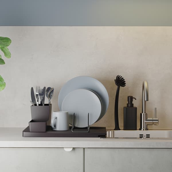 Czarna suszarka na naczynia z szaroniebieskimi talerzami i sztućcami ustawiona obok wpuszczanego zlewu ze stali nierdzewnej.