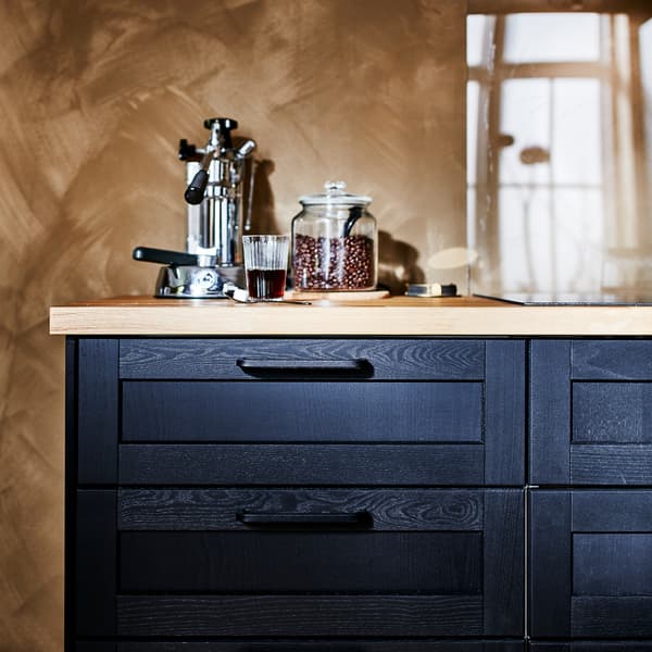 Czarna szafka stojąca z czarnymi frontami szuflad i drewnianym blatem. Na blacie stoi zaparzacz do kawy i słoik z ziarnami kawy.