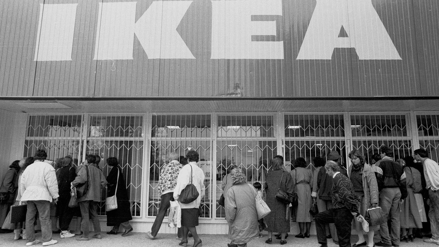 Czarno-białe zdjęcie pierwszego sklepu IKEA w Polsce. Ludzie stoją w kolejce do blaszanego budynku, na którym widnieje wielki napis "IKEA".