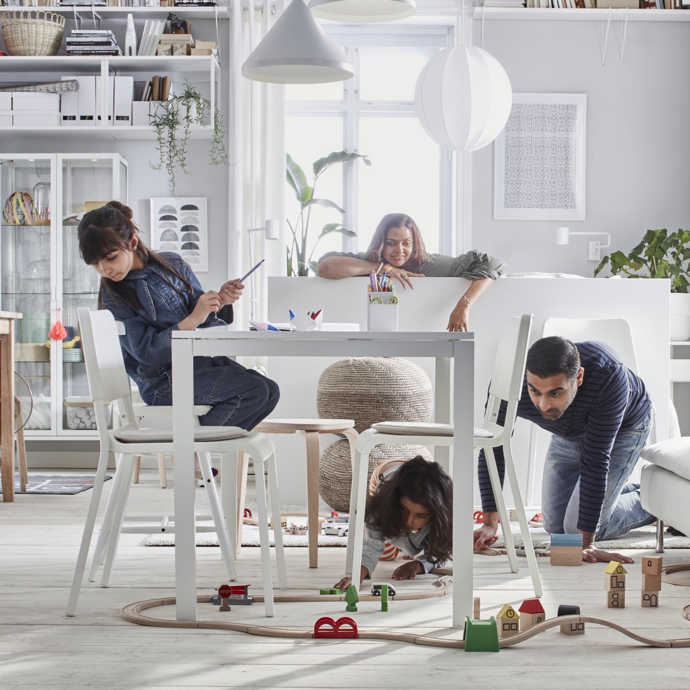 ダイニングテーブルの下で大人が膝をついて子どもと遊んでいる。その後ろに大人がもう1人いる。