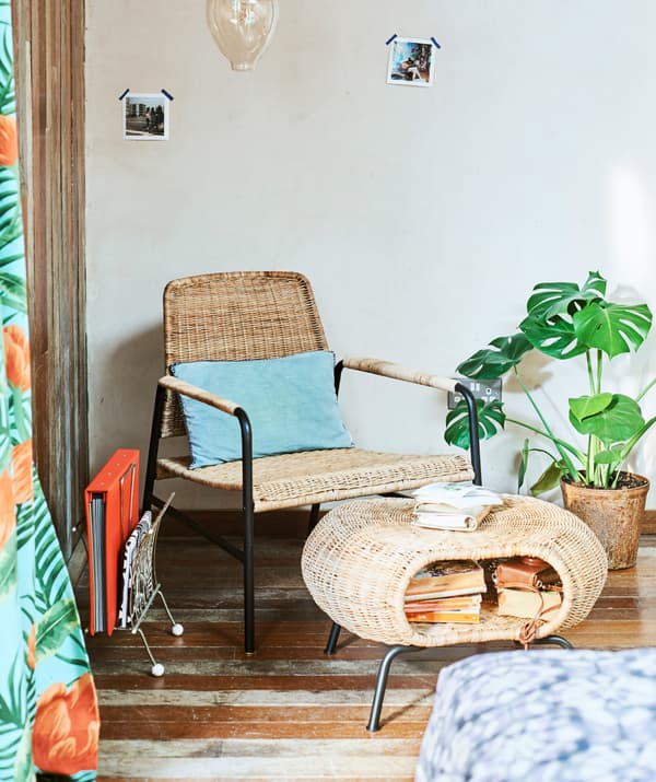 Dans l’angle d’une pièce, un fauteuil en rotin et un repose-pieds en rotin avec un rangement plein de livres; suspendue au-dessus de l’ensemble, une ampoule décorative.