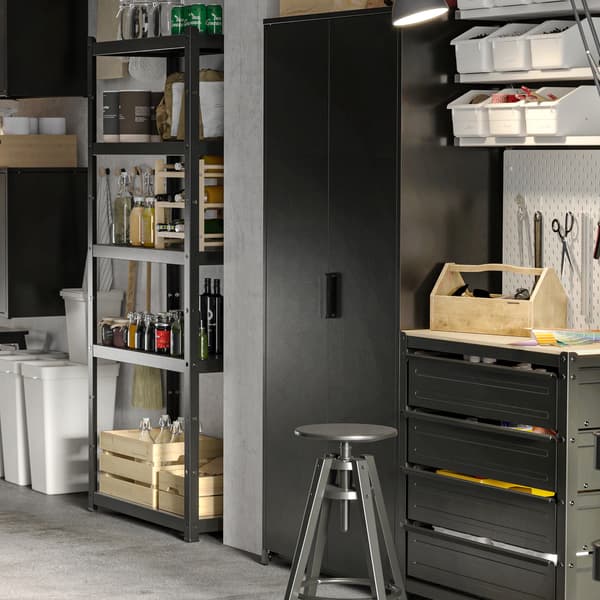 Dans un garage, armoires et rangements ouverts et avec portes en noir. Sur les tablettes, boîtes, flacons et bouteilles. Au sol, bacs de recyclage.