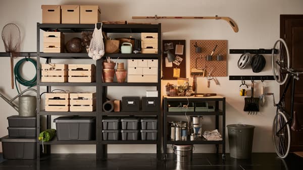Dans un garage, des étagères BROR noires sont fixées au mur et sont remplies de boîtes en bois, en carton et en plastique de différentes dimensions.