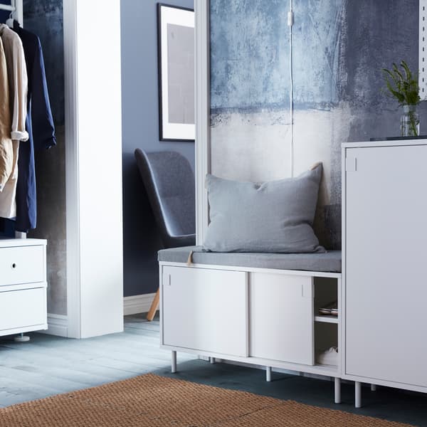 Dans un hall d’entrée, un banc avec rangement MACKAPÄR garni d’un coussin gris, à côté d’une armoire à chaussures MACKAPÄR sur laquelle est posé un vase.