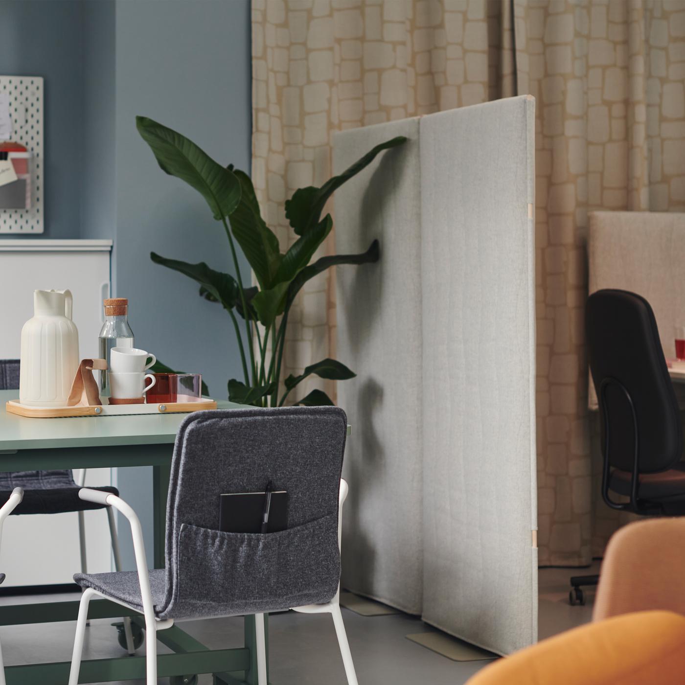 Dans un petit bureau aux murs bleus et aux rideaux beiges, chaise LÄKTARE grise avec poche, et grande plante verte dans un coin. 