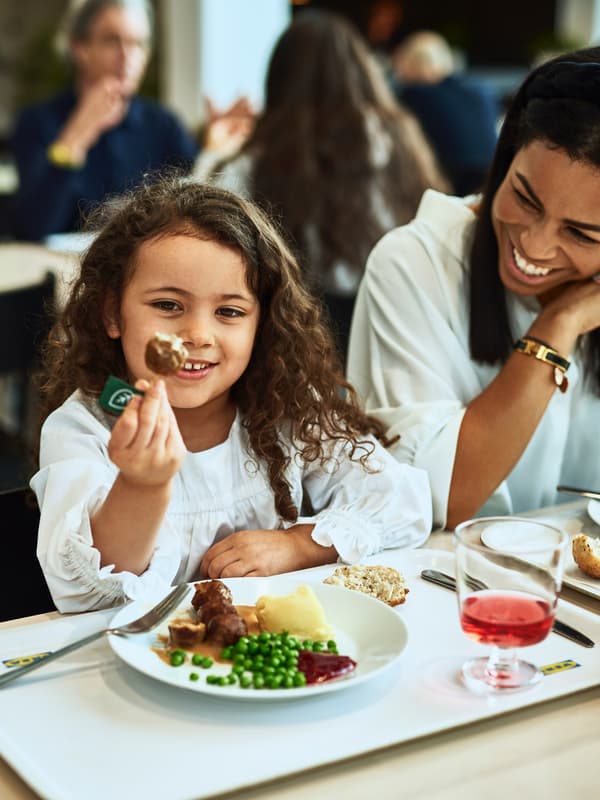 Dans un restaurant IKEA, une petite fille déguste un plat de boulettes végétales, de purée de pommes de terre et de petits pois sous le regard de sa mère.