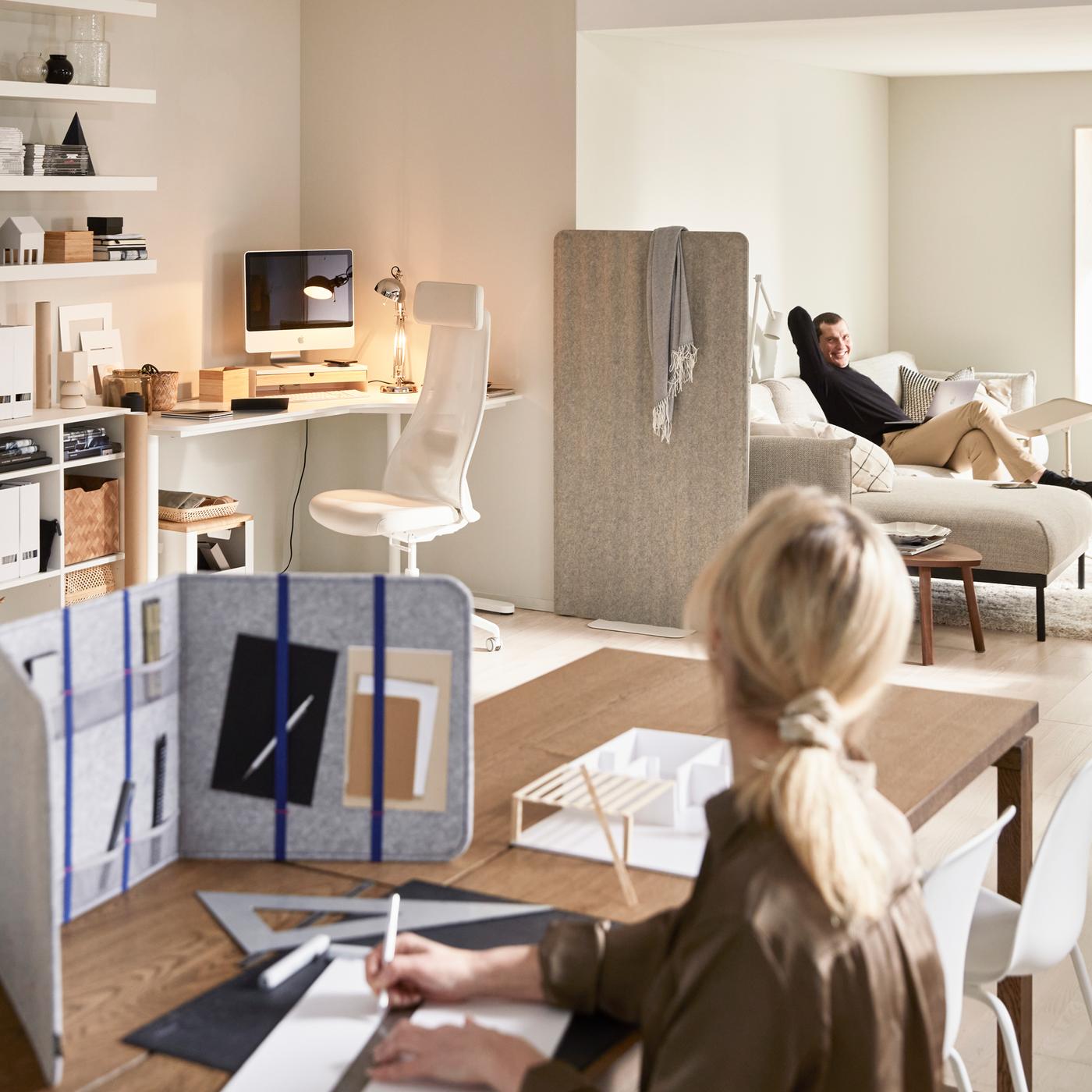 Dans un salon, une femme utilisant un séparateur de bureau ÖVNING pour étudier, un bureau BEKANT dans un coin et un homme assis sur un canapé.