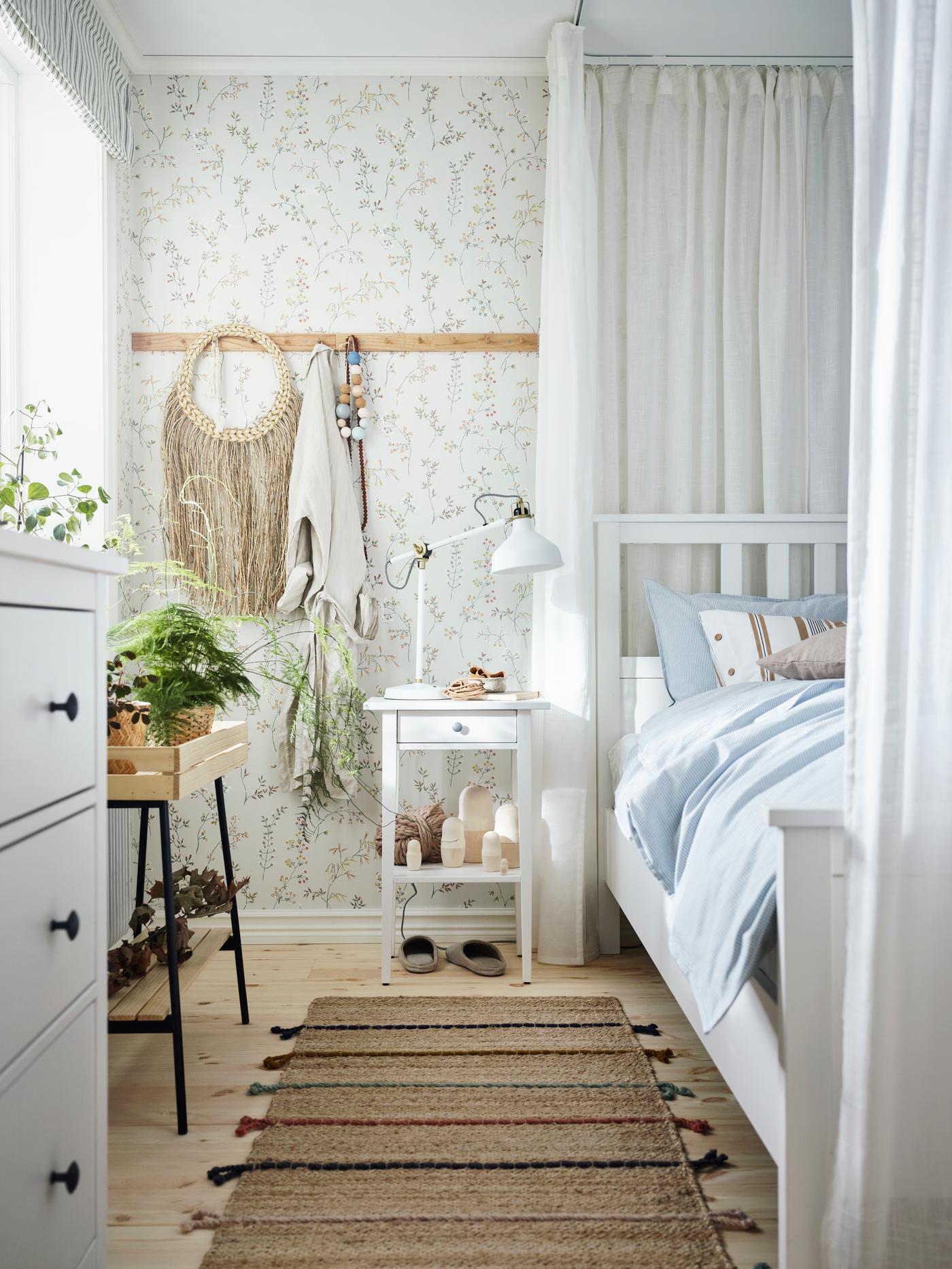Dans une chambre ensoleillée, un lit HEMNES blanc et une table de chevet avec une lampe de travail RANARP, des plantes et des objets décoratifs naturels.