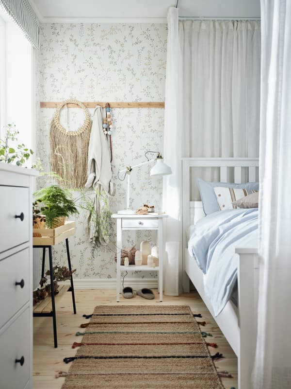 Dans une chambre ensoleillée, un lit HEMNES blanc et une table de chevet avec une lampe de travail RANARP, des plantes et des objets décoratifs naturels.