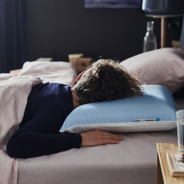Dans une chambre sombre, une personne en pyjama bleu dort sur le ventre, la tête posée sur un oreiller ergonomique KVARNVEN.