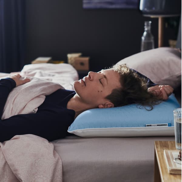 Dans une chambre sombre, une personne en pyjama bleu dort sur le dos, la tête posée sur un oreiller ergonomique KVARNVEN.