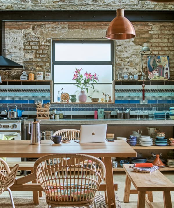 Dans une cuisine de style industriel, une table en bois avec des bancs et des chaises en rotin, des carrelages bleus au mur et de la vaisselle empilée sur des étagères ouvertes.