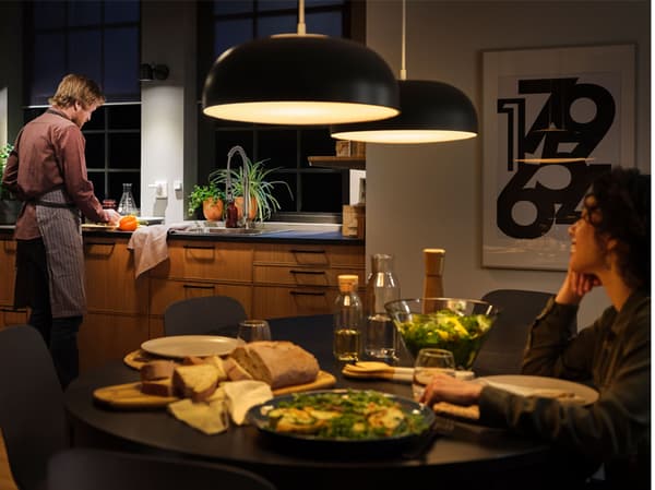 Dans une cuisine dotée d’un éclairage connecté, dont de grandes suspensions au-dessus de la table, une femme est assise à la table mise et un homme est debout devant l’évier.