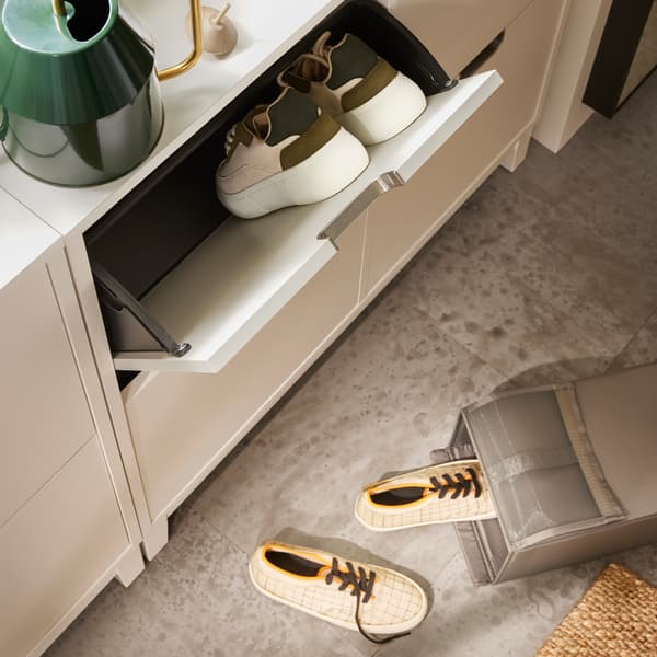 Dans une pièce, armoire à chaussures blanche fermée. Un tiroir est ouvert et révèle des baskets. Arrosoir vert sur le dessus de l’armoire.