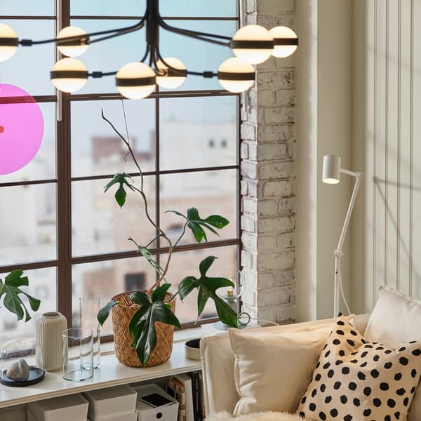 Dans une pièce bien éclairée, un lustre STORSLINGA noir et blanc et un lampadaire NYMÅNE blanc à côté d’un canapé blanc avec un coussin.