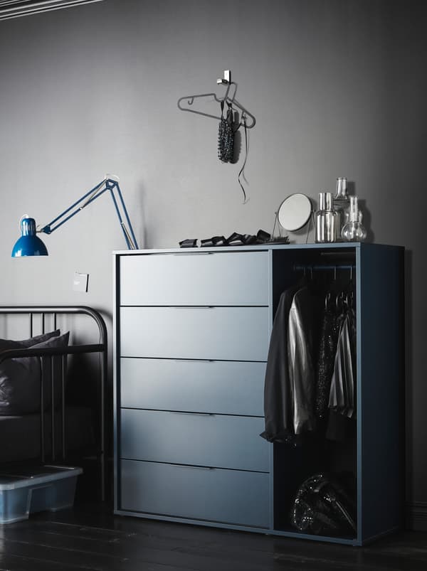 Dans une pièce d’allure minimaliste aux murs gris, des vêtements sont suspendus sur la tringle d’une commode NORDMELA foncée, alors que des décorations et un miroir sont posés sur le dessus.