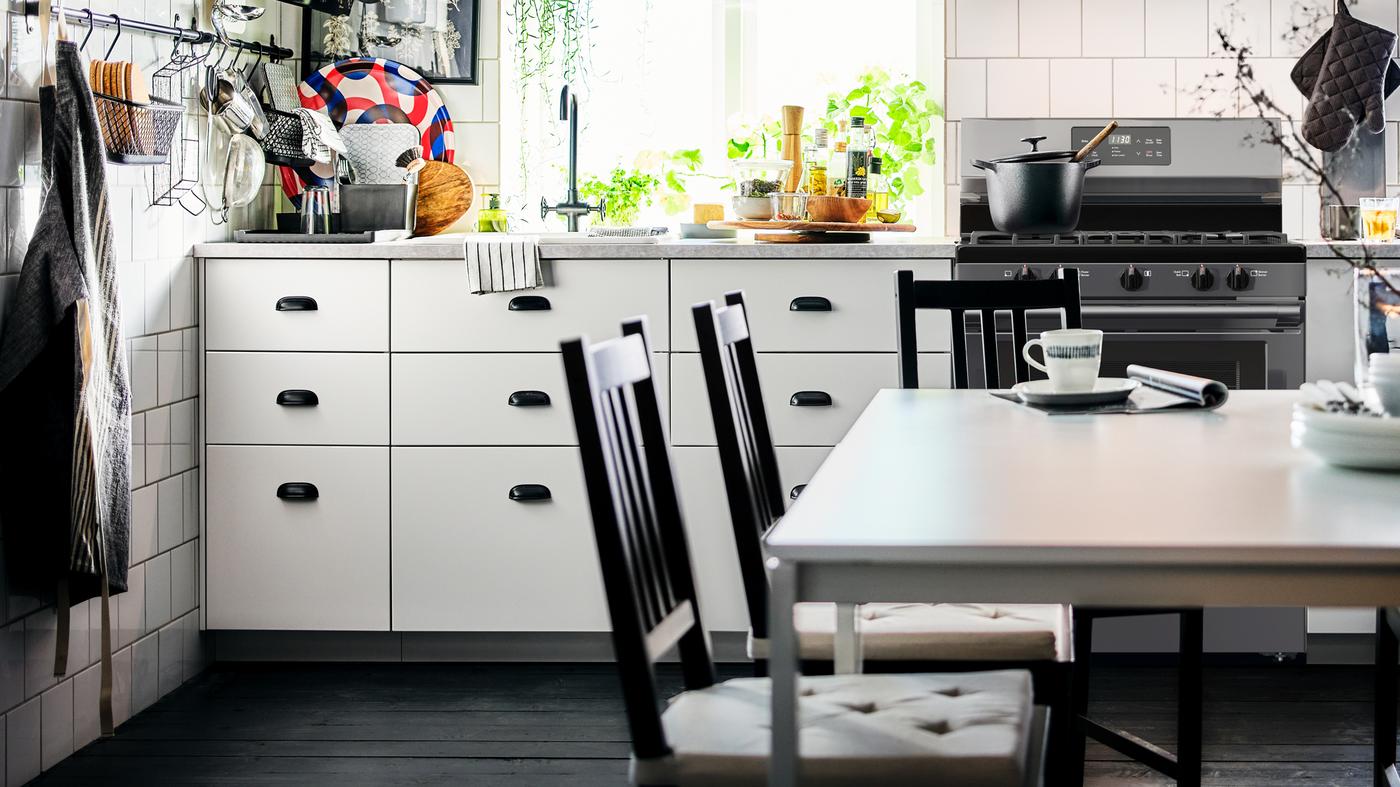 Dapur berwarna hitam dan putih yang mempunyai kabinet dasar berwarna putih dan ruang makan yang berpusat di sekitar dua meja MELLTORP berwarna putih.